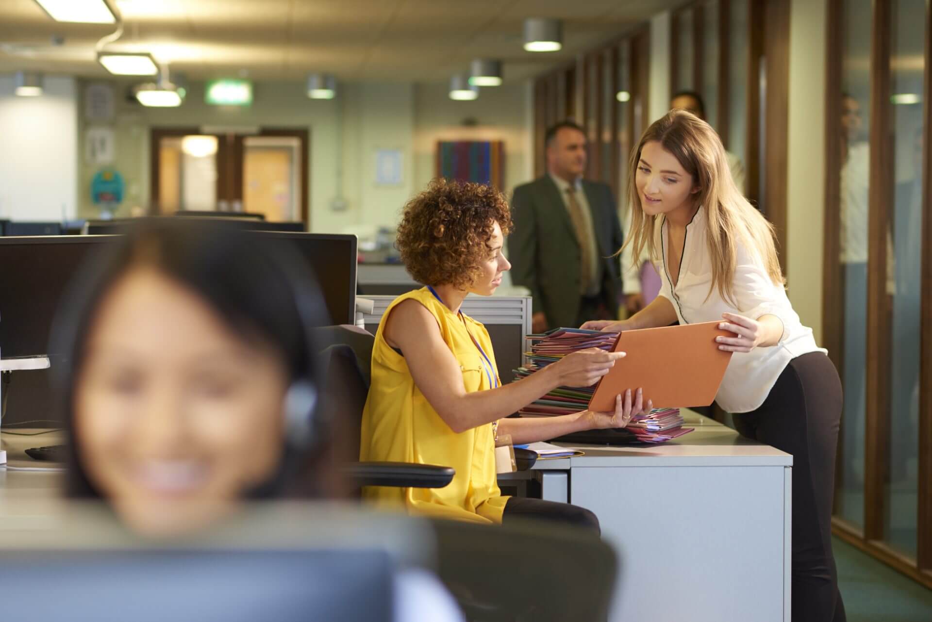 a young office junior takes a file from her supervisor for filing in a large open plan office . Co-workers can be seen defocussed in the foreground and background