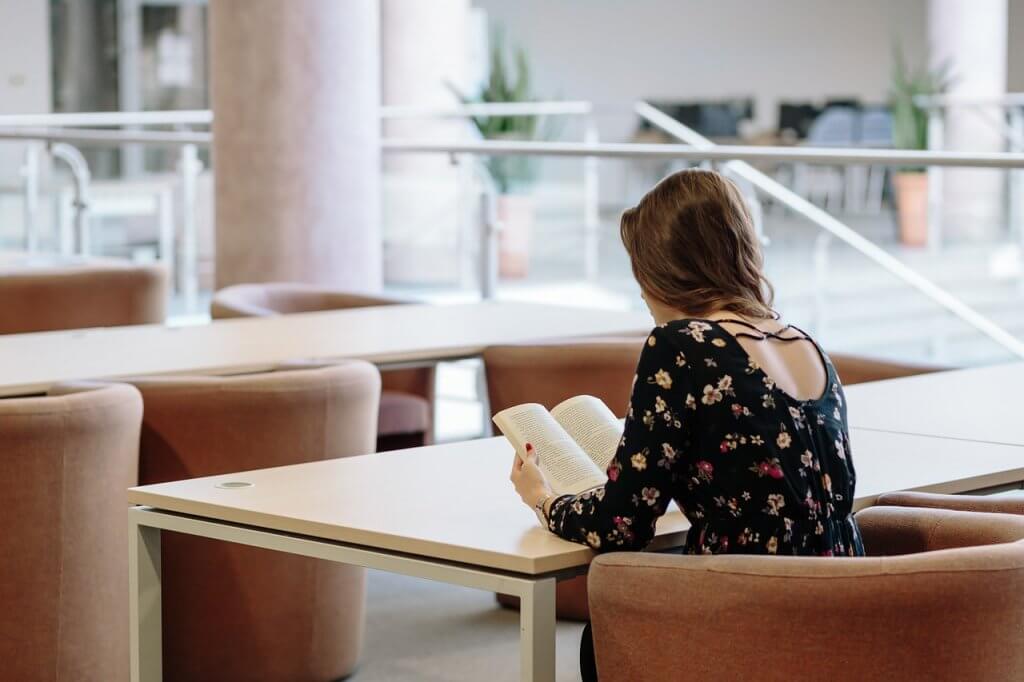 Student sitting on a round couch in an open space, leaning on a desk and reading a book