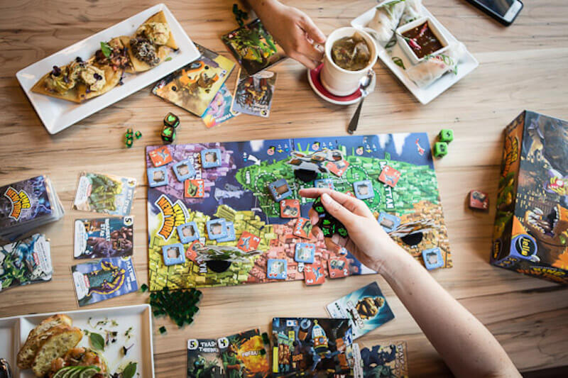 Board game in middle of table with an array of food and drinks surrounding 