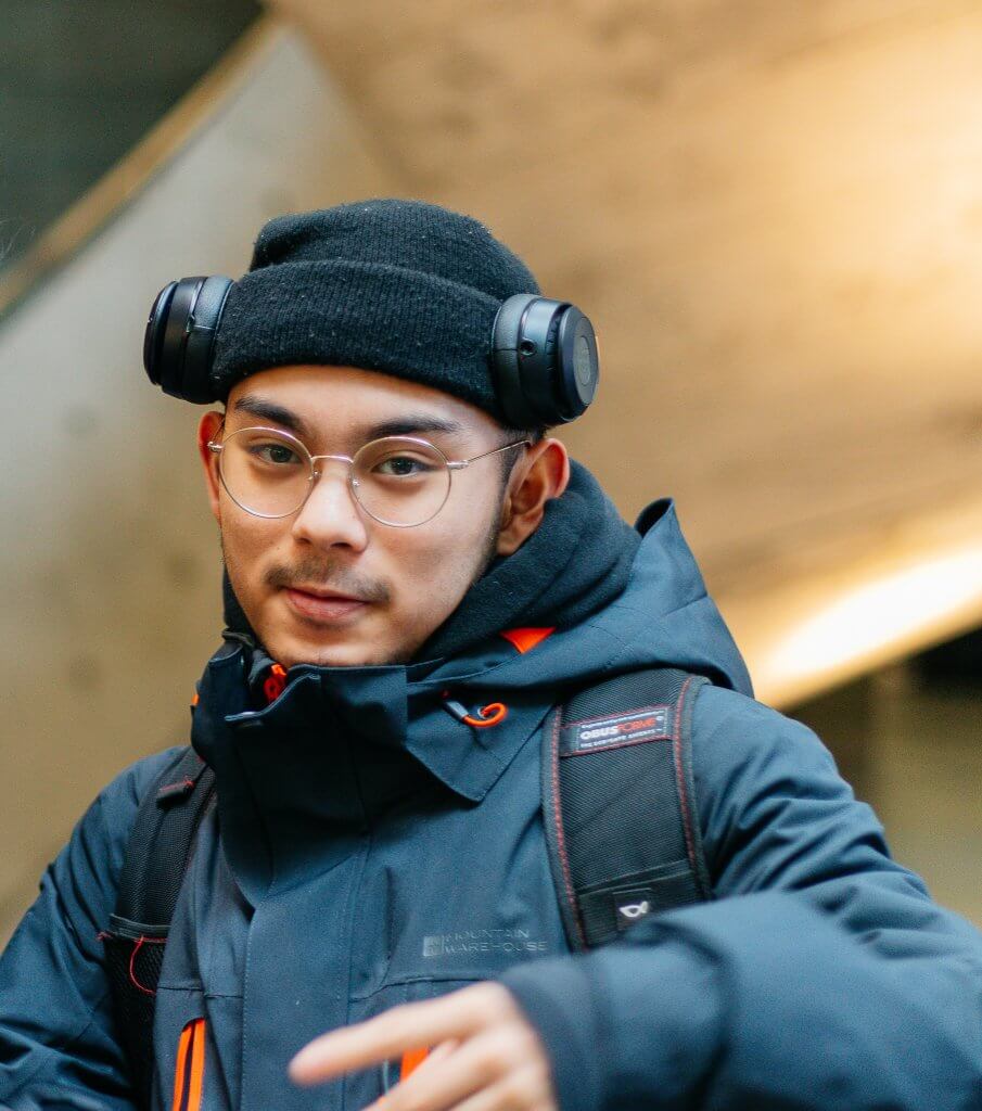 man wearing jacket, beanie with headphones, and glasses, while smirking.