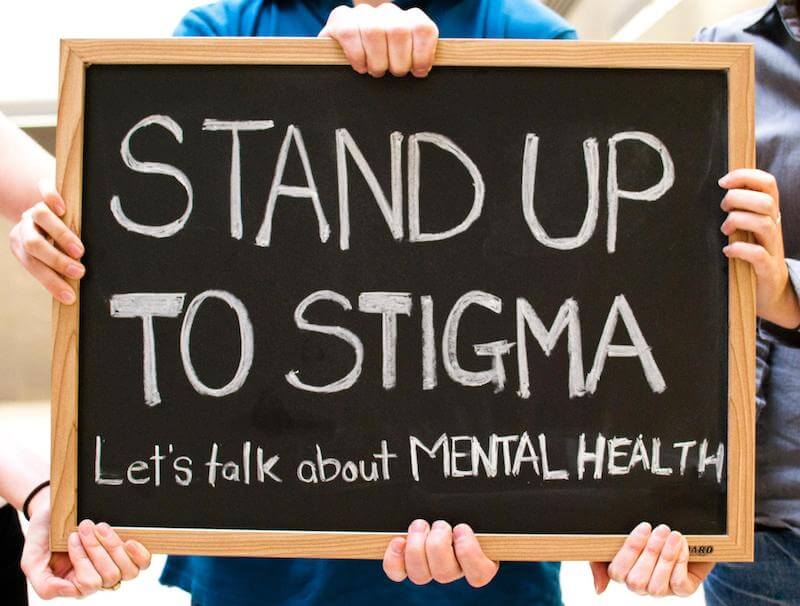 Hands holding up a blackboard sign that reads Stand up to stigma. Let's talk about mental health.