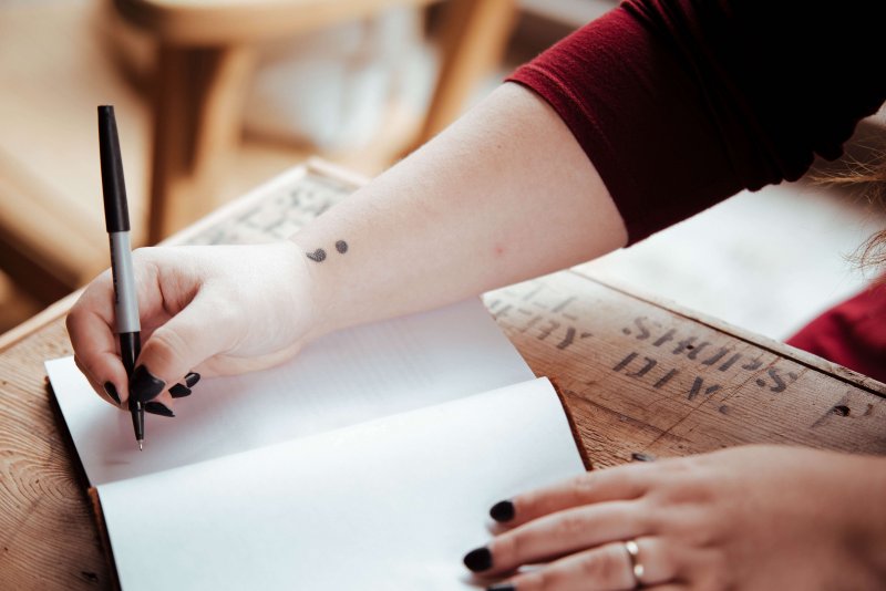 girl writing in notebook with semi colon tattoo on arm