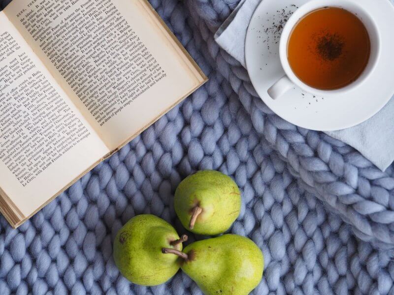 Open novel, 3 pears, and cup of tea on top of knitted blanket