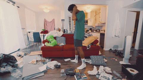 A young man wearing a green shirt and black basketball shorts appears confused as he searches through a messy living room.