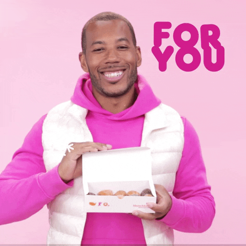 A man wearing a bright pink hooded sweatshirt and puffy white vest smiles while holding out a box of doughnuts to the camera. The words "For you" appear in the upper right corner.