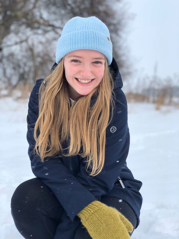 Second year Humber College Food and nutrition management student Amy Hasson kneels in the snow while wearing a tuque and a winter jacket and smiles.