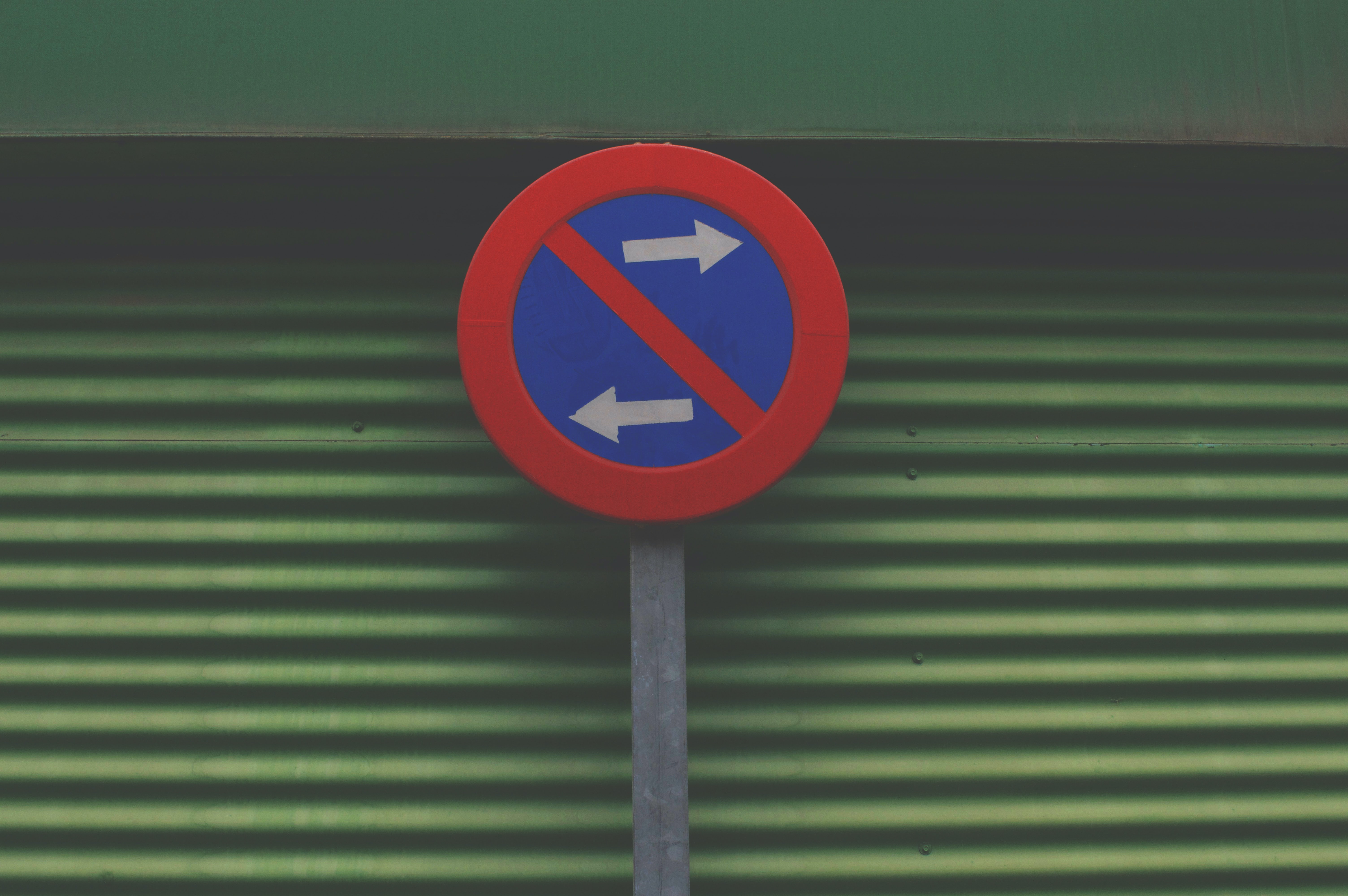 A circular sign stands against a textured army green wall. The sign is royal blue with a red border and two white arrows, one pointing left, the other pointing right.