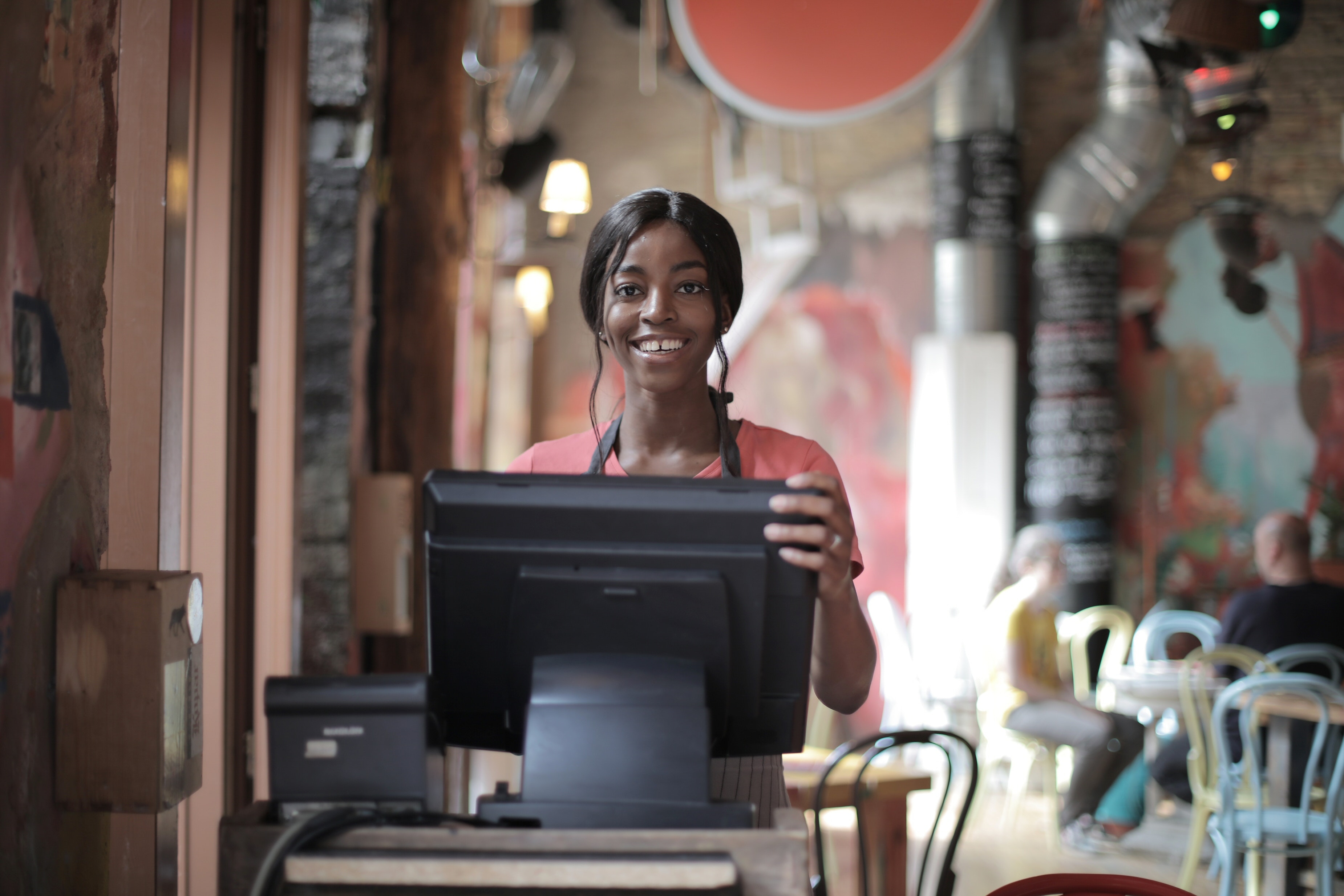 A young woman in a pink shirt and a black apron stands behind a cash register, smiling. She has dark hair that is gathered in a low ponytail with two strands dangling in front of her face.