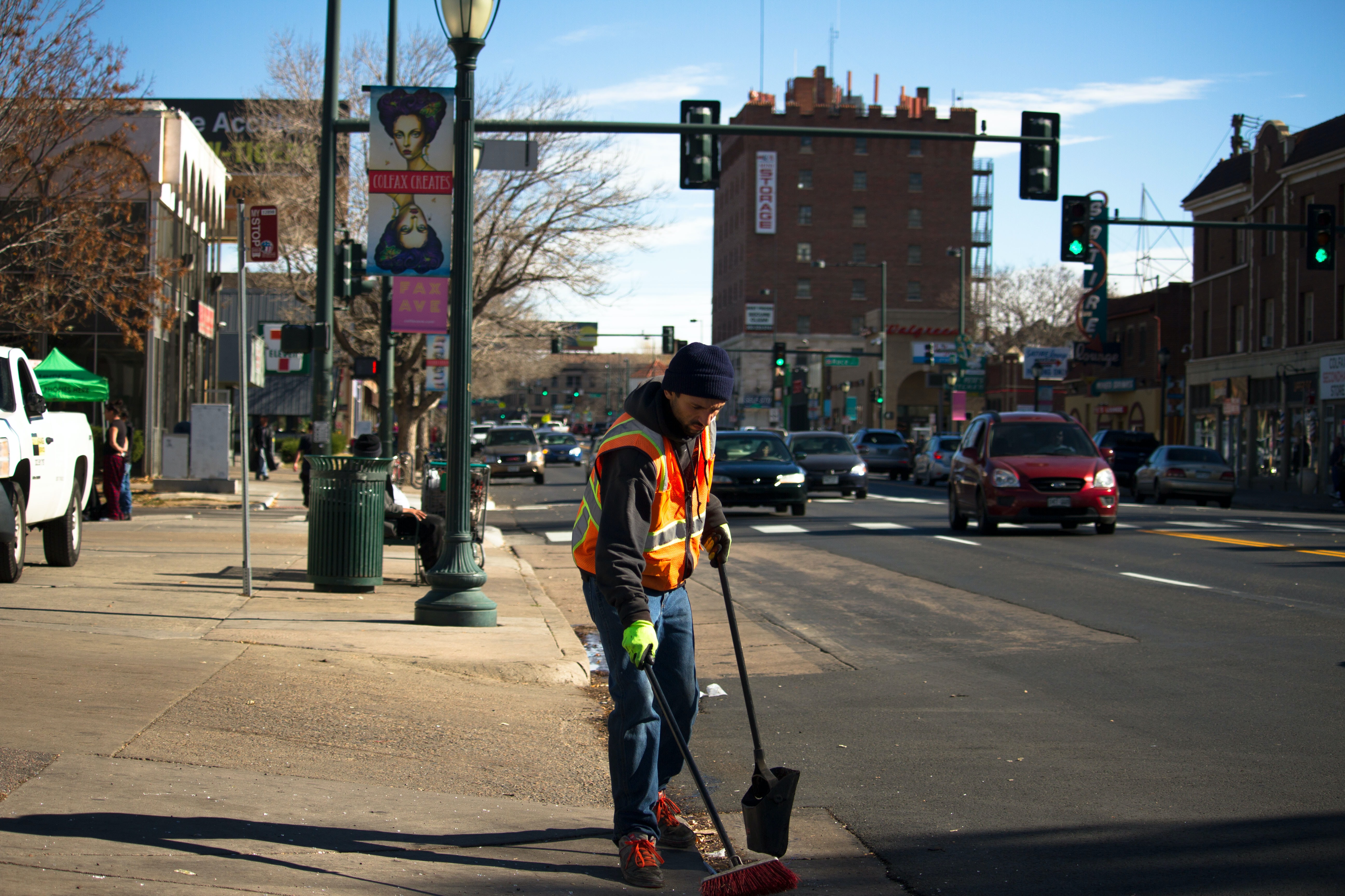 A young man wearing a navy blue toque, dark blue bootcut jeans, neon yellow gloves, a grey sweater, and a bright orange reflective vest sweeps a city street with a broom and dustpan.