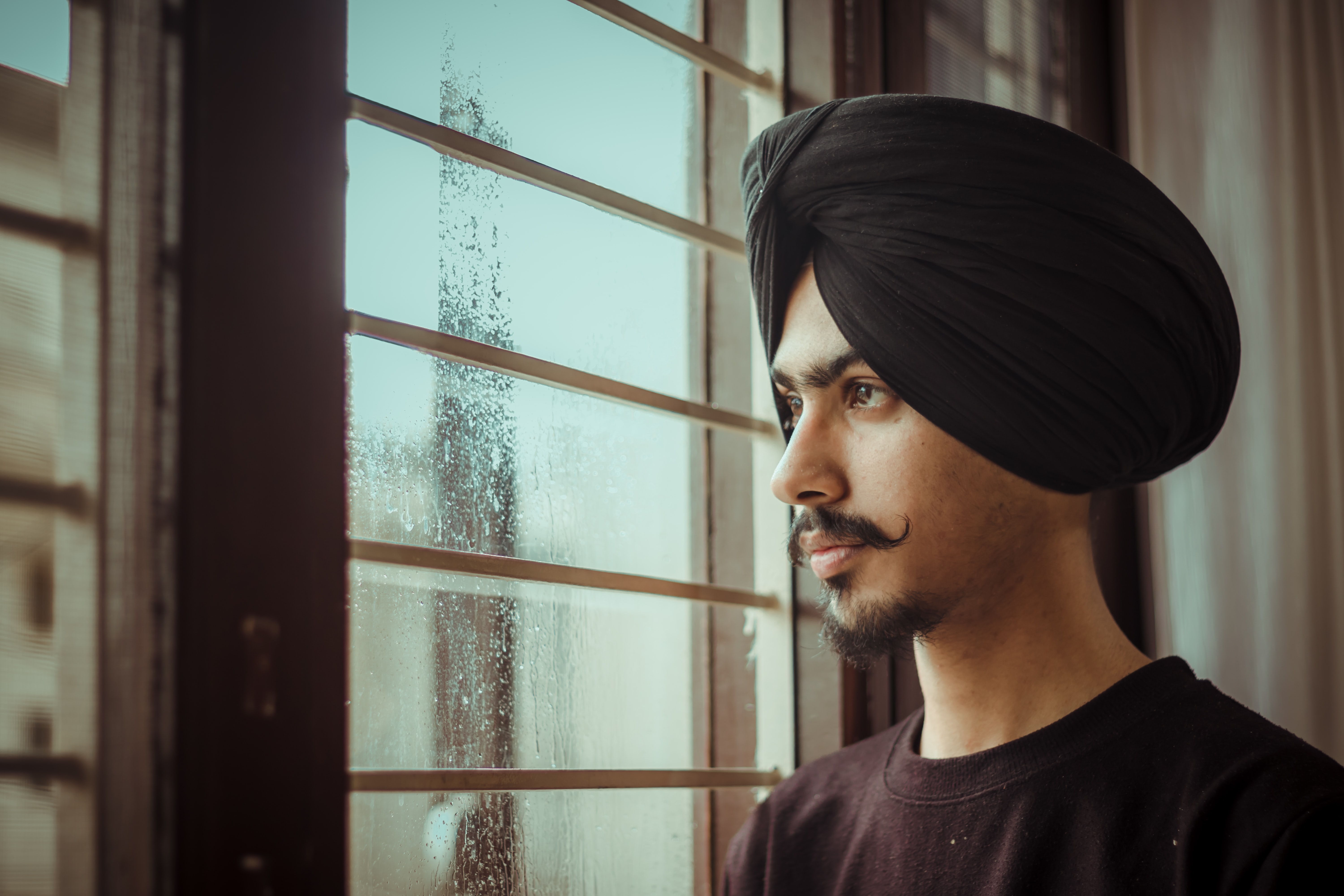 A young man with a moustache wearing a black turban stands next to a window, staring outside solemnly.
