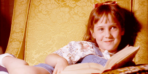 Mara Wilson as Matilda Wormwood in "Matilda" (1996) sits on an armchair reading a book and giggles. 