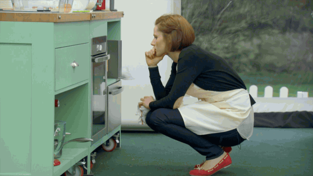 A chef with red hair wearing a white apron, black shirt, black pants and red shoes squats in front of an oven, watching as her bread bakes.