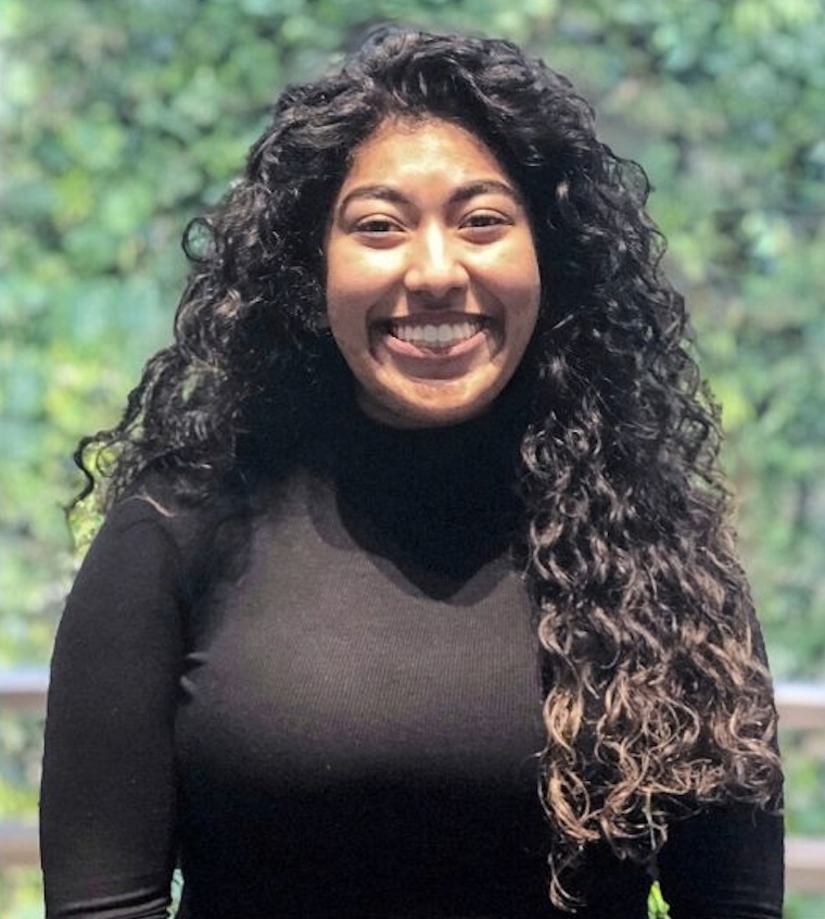 IGNITE 2020-2021 Board of Directors member Megan Roopnarine smiles at the camera while wearing a black turtleneck sweater. She has long, dark, curly hair.
