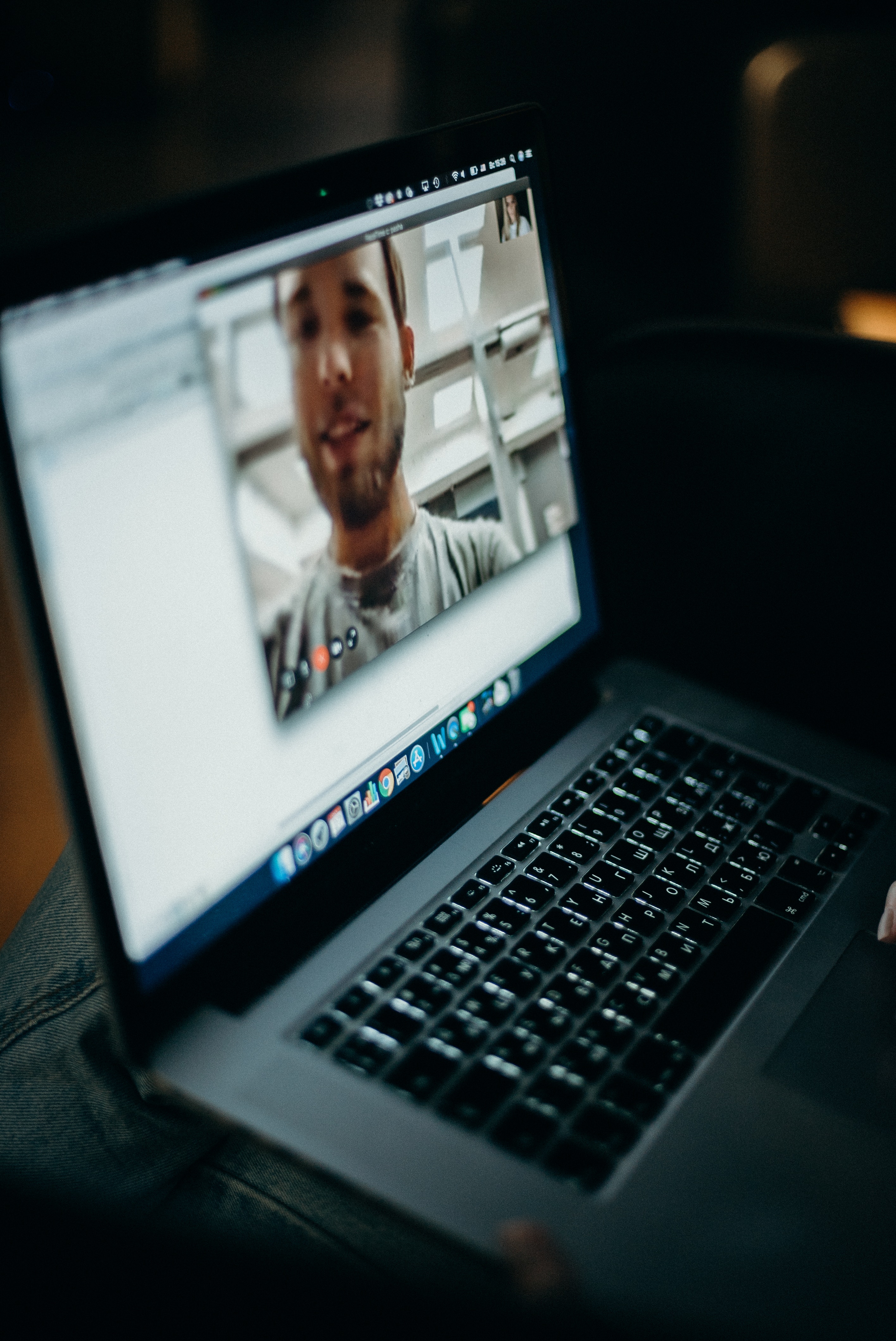 A silver MacBook with black keys displays a video call with a young man with short, dark hair.
