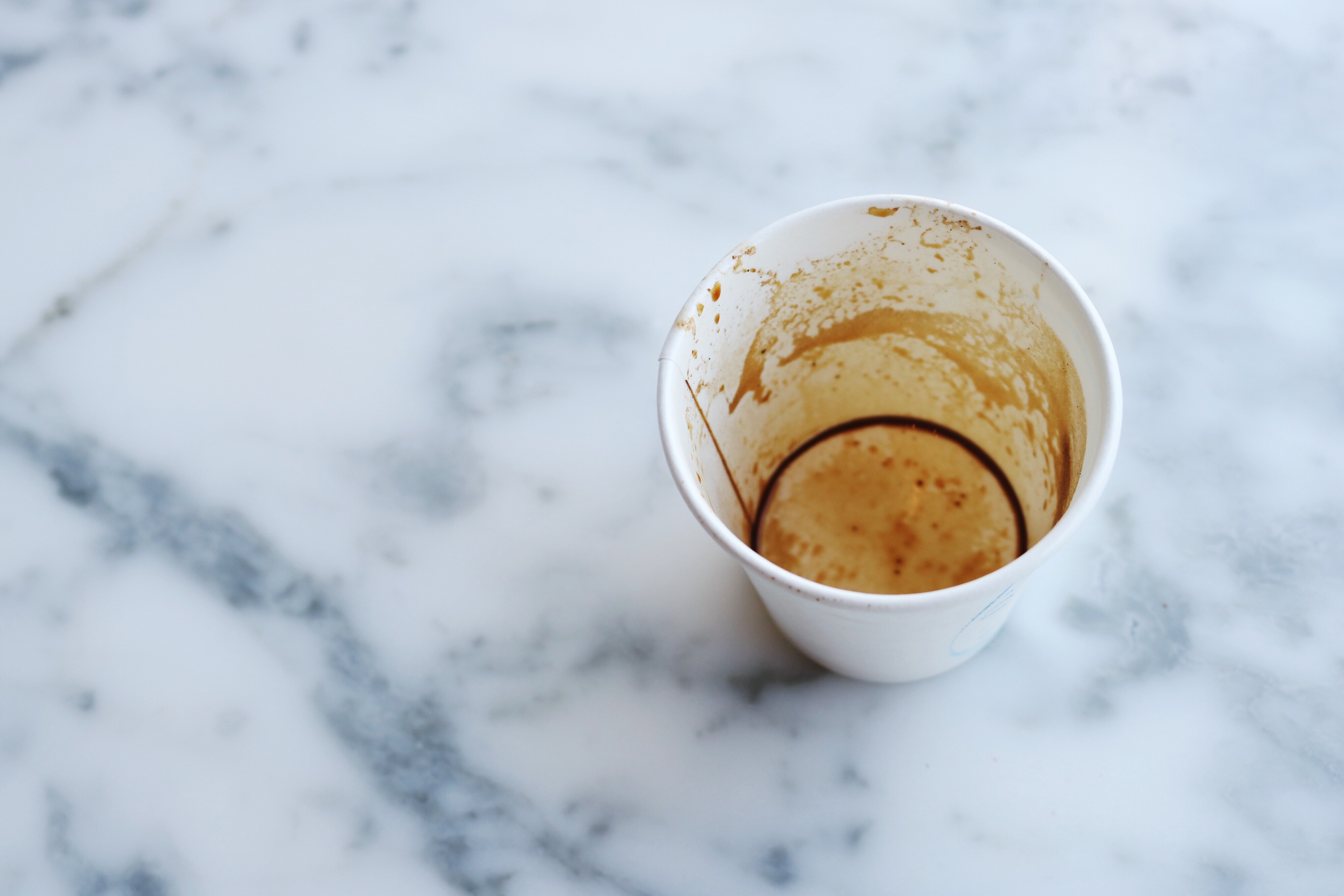 An empty white paper coffee cup sits atop a white and grey marble counter. The inside of the cup is stained with remnants of coffee.