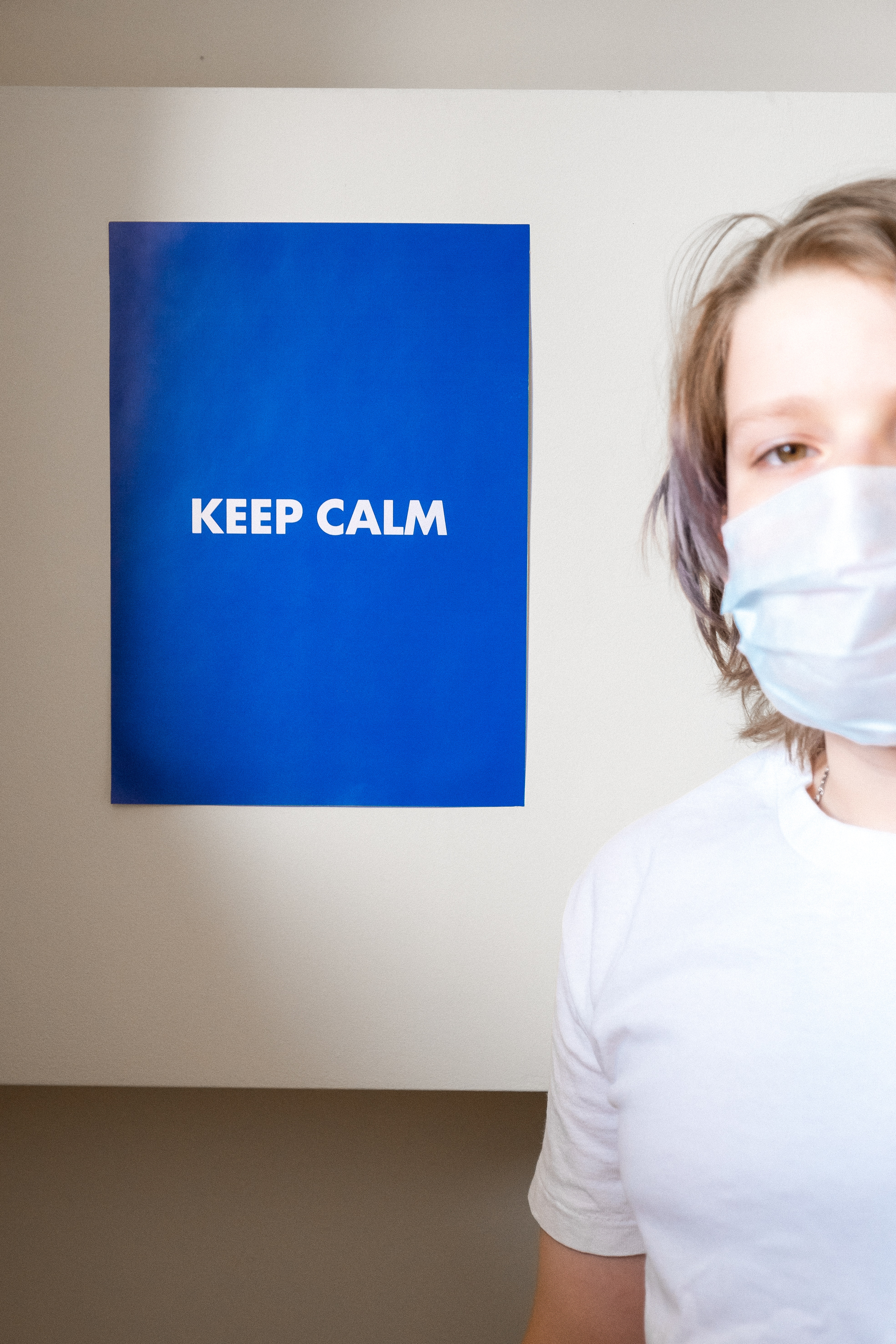 A young woman with chin-length shaggy blonde hair wears a white shirt and stands next to a royal blue sign with the words "Keep calm" printed on it in white block letters. The woman wears a cloth face mask over her mouth.