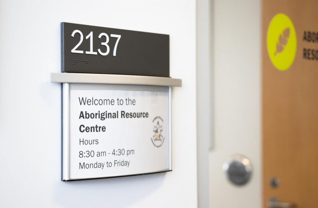 A white, slightly ajar door dons a small black sign with the number "2137" printed on it in white. Below the black sign is a slightly larger sign that reads, "Welcome to the Aboriginal Resource Centre. Hours: 8:30 a.m.-4:30 p.m. Monday to Friday."