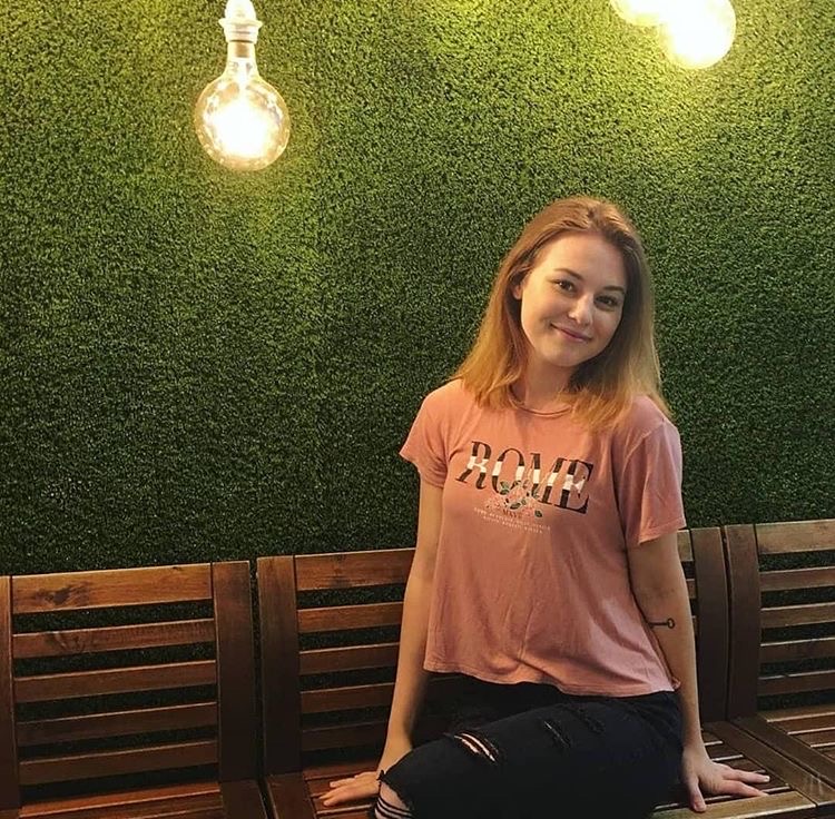 University of Guelph-Humber Public Relations Student Laik Sweeney wears a pink T-shirt while standing in front of a green wall. She smiles at the camera.