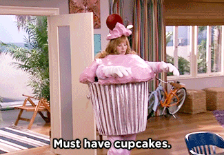 A woman dressed in a 3D pink cupcake costume says, "Must have cupcakes".