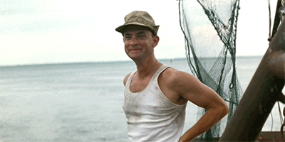Tom Hanks as Forrest Gump wears a dirty white tank top and a ballcap. He smiles and waves dramatically.