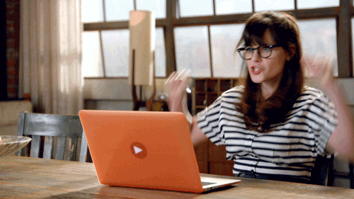 Zooey Deschanel silently cheers while sitting at a desk in front of an orange laptop.