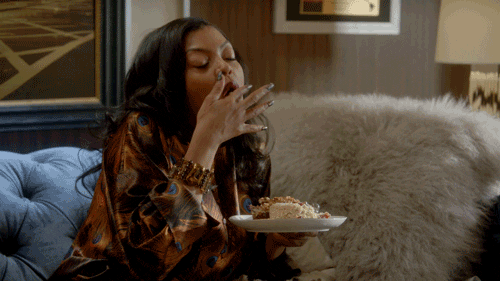 Actress Taraji P. Henson sits on a sofa holding a plate of cake. She takes a bite and licks her fingers.