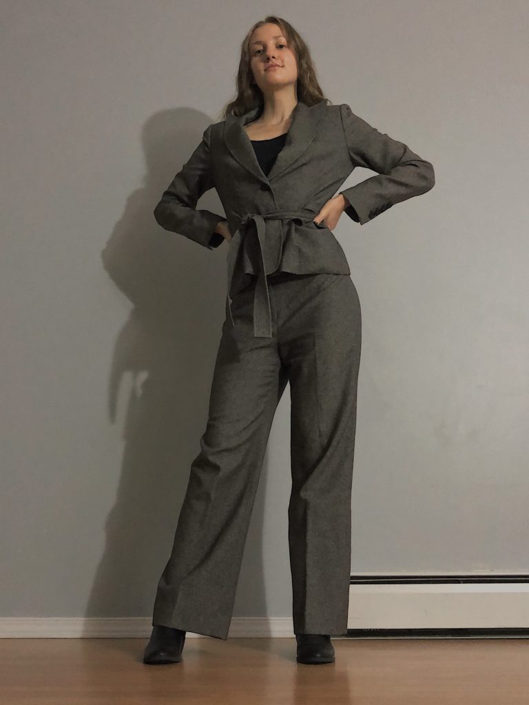 Gabby wears a grey pantsuit with her hands on her hips.  