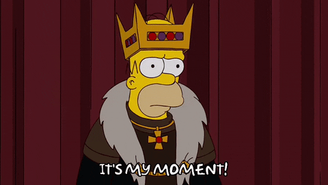Homer Simpson, wearing a crown, says "It's my moment!"