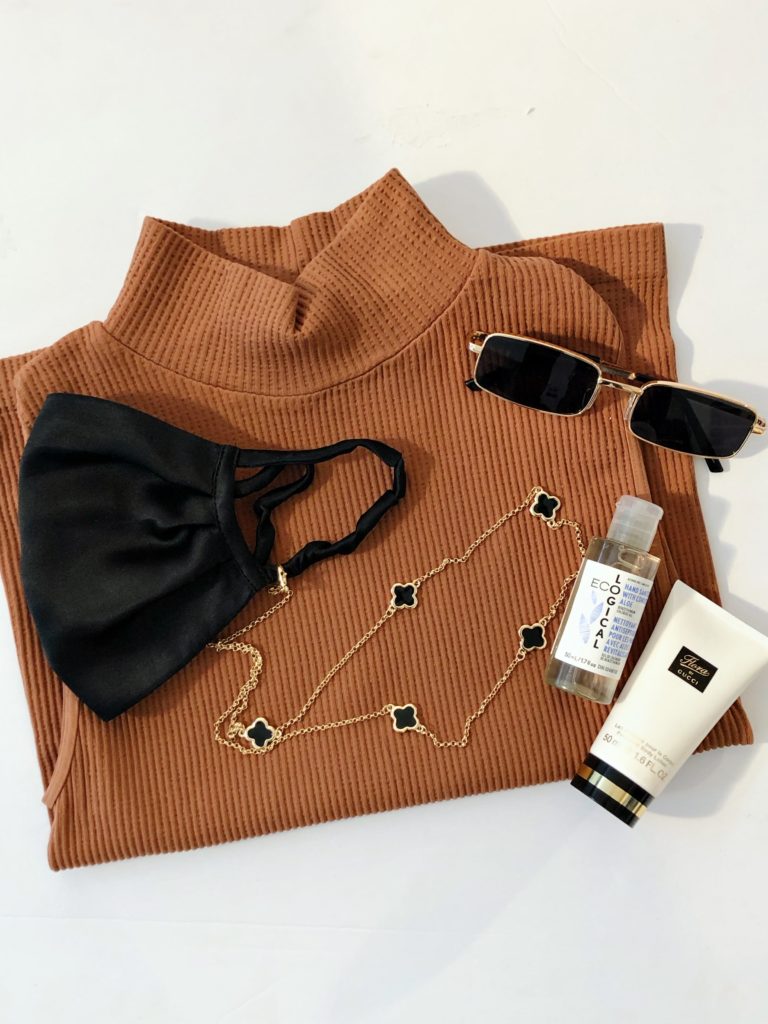 Brown turtleneck shirt against a white background with a black face mask, gold and black chain, and sunglasses laying on top of it.