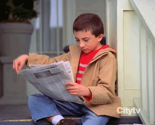 A boy reading a newspaper putting a finger up to person out frame to tell them to wait.