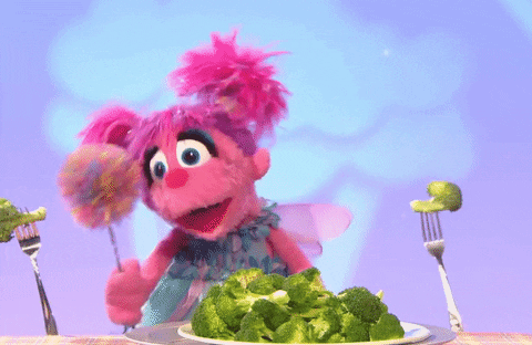A pink muppet dancing next to a plate of broccoli.