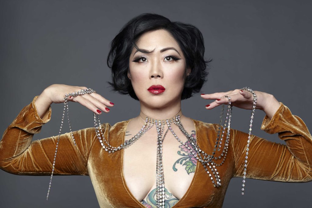 Margaret Cho posing with her hands raised to chin level against a grey background.