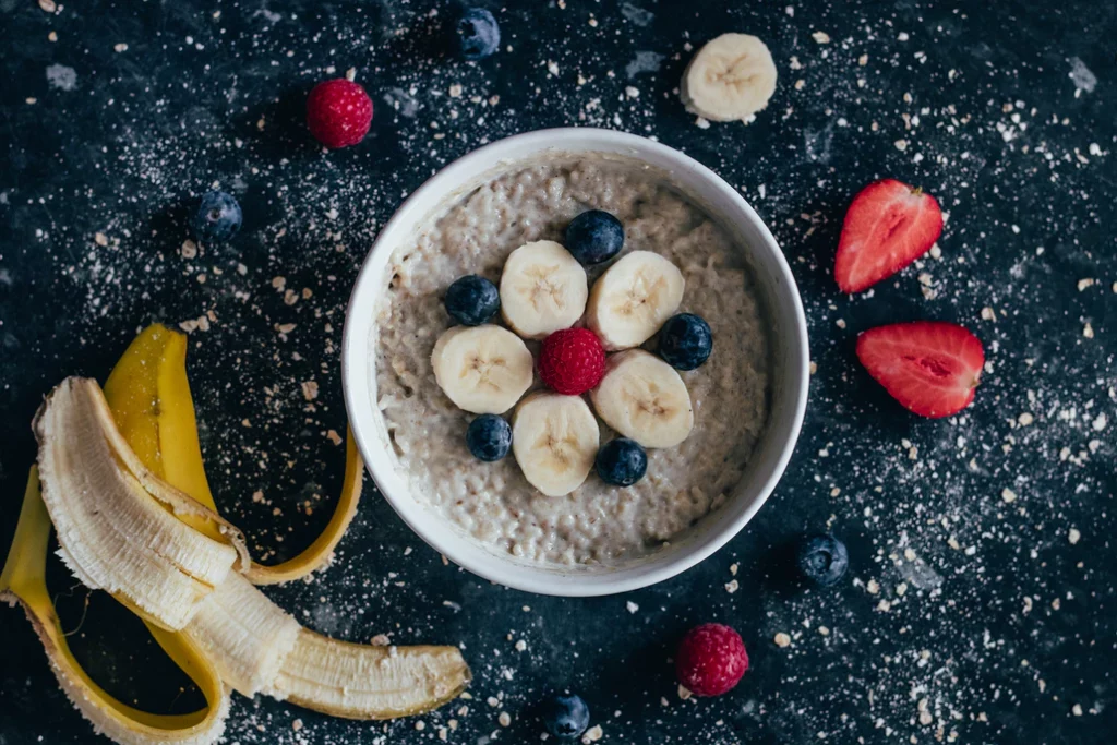 Bowl of oatmeal with bananas and blueberries arranged in the middle and various fruits surrounding the bowl.