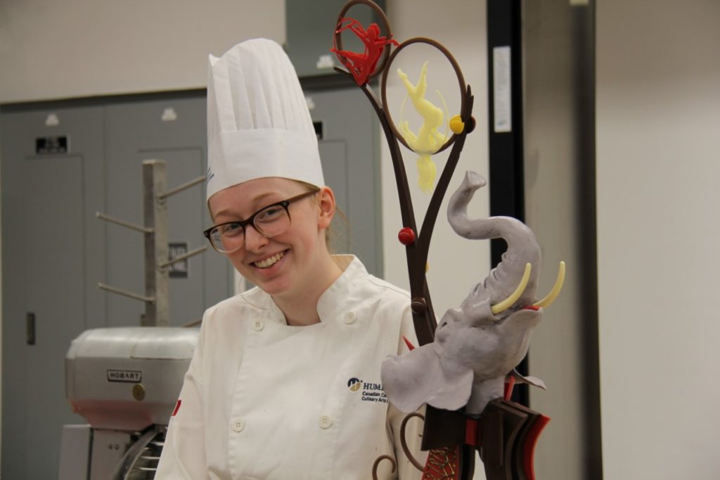 Emma is dressed in Humber's s chef uniform. She is standing next to one of her baking creations.