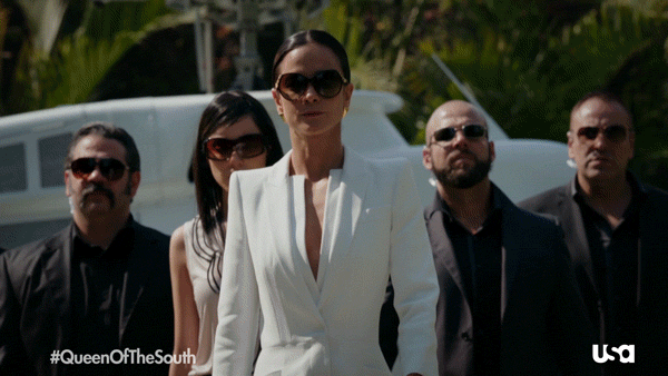 Teresa from the series "Queen of the South" is dressed in a white dress and walking with her bodyguards behind her.