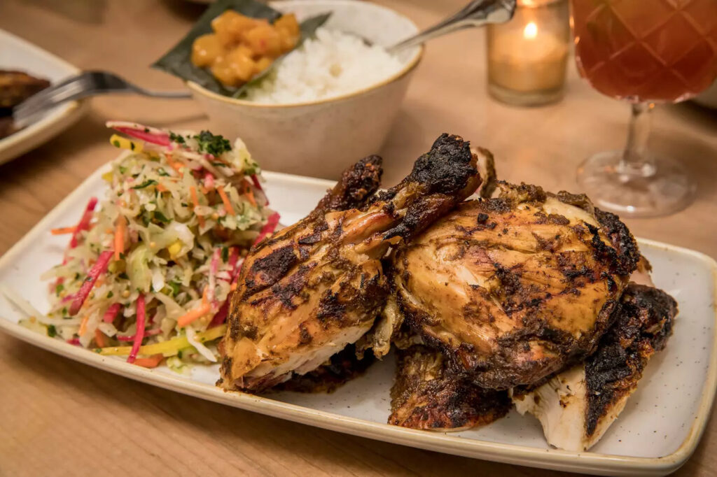A serving of jerk chicken and salad on a white plate. Behing the jerk chicken, there is a serving of rice.