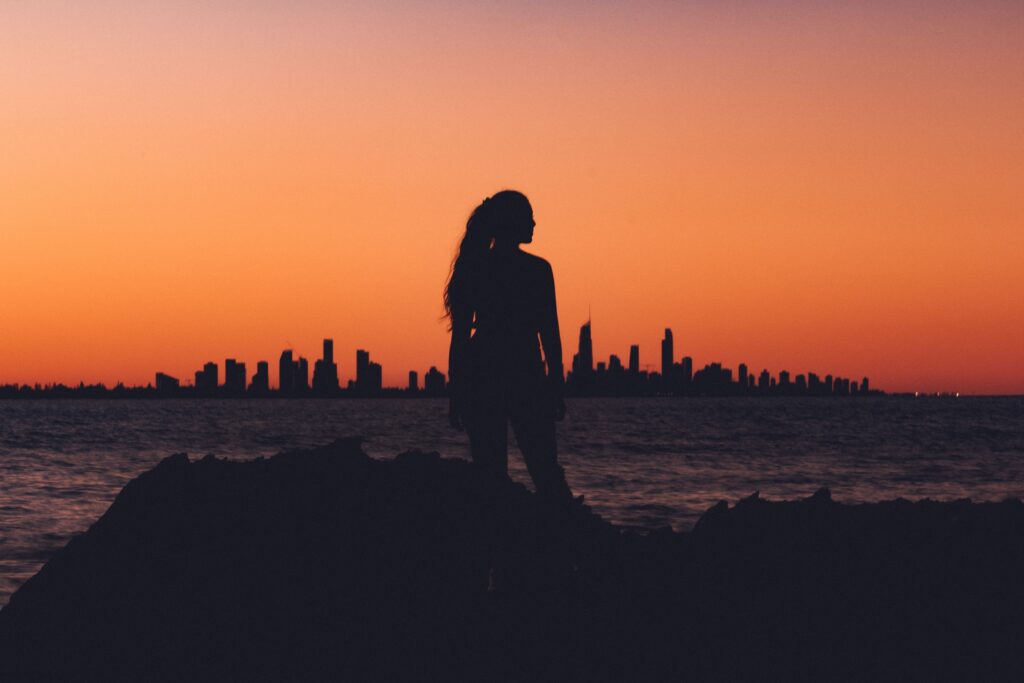 Silhouette of a woman by the water with a city skyline in the distance.