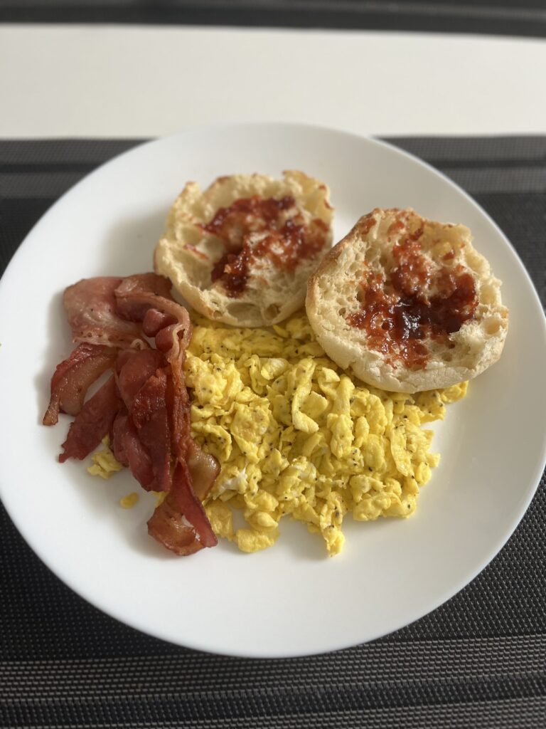 A plate of scrambled eggs and bacon served with English muffins and guava jam.