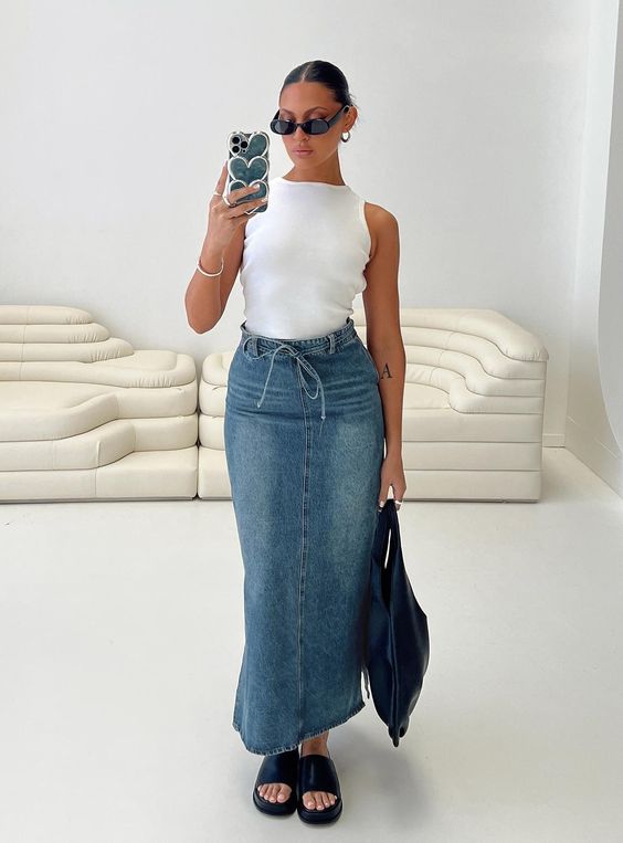 A girl is wearing a maxi denim skirt. She pairs it with a white tee, sunglasses and platform sandals. 