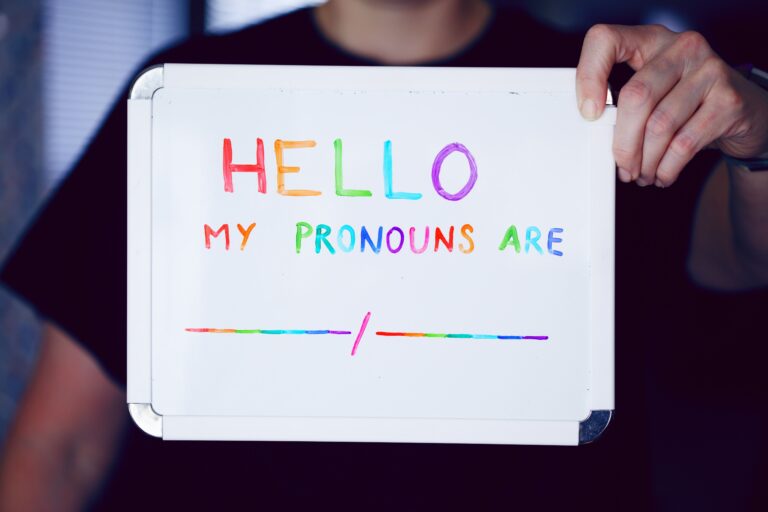 Picture of a person holding a whiteboard with the sentence “Hello my pronouns are” in pride colours.