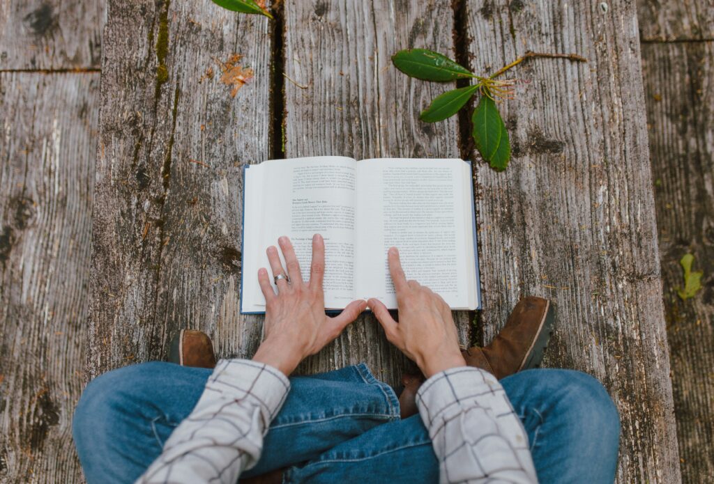 Picture of a person reading a book outdoors on top of a wood bench.