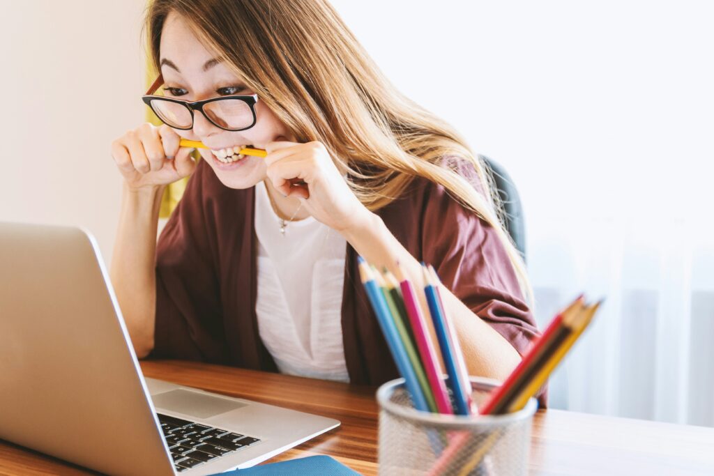 A woman biting a pencil while sitting on a chair in front of a computer.