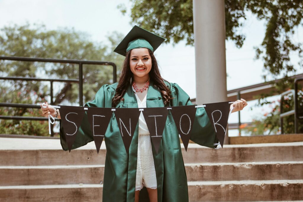 A woman wearing a graduation regalia and holding a black sign that reads “senior.”