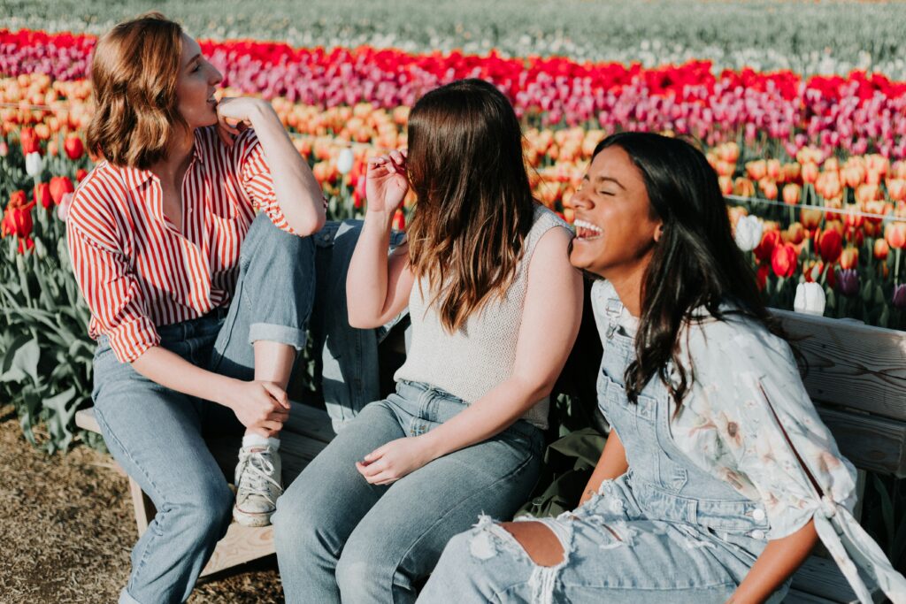 Three women sitting on a wooden bench by a tulip flower field and they are laughing.