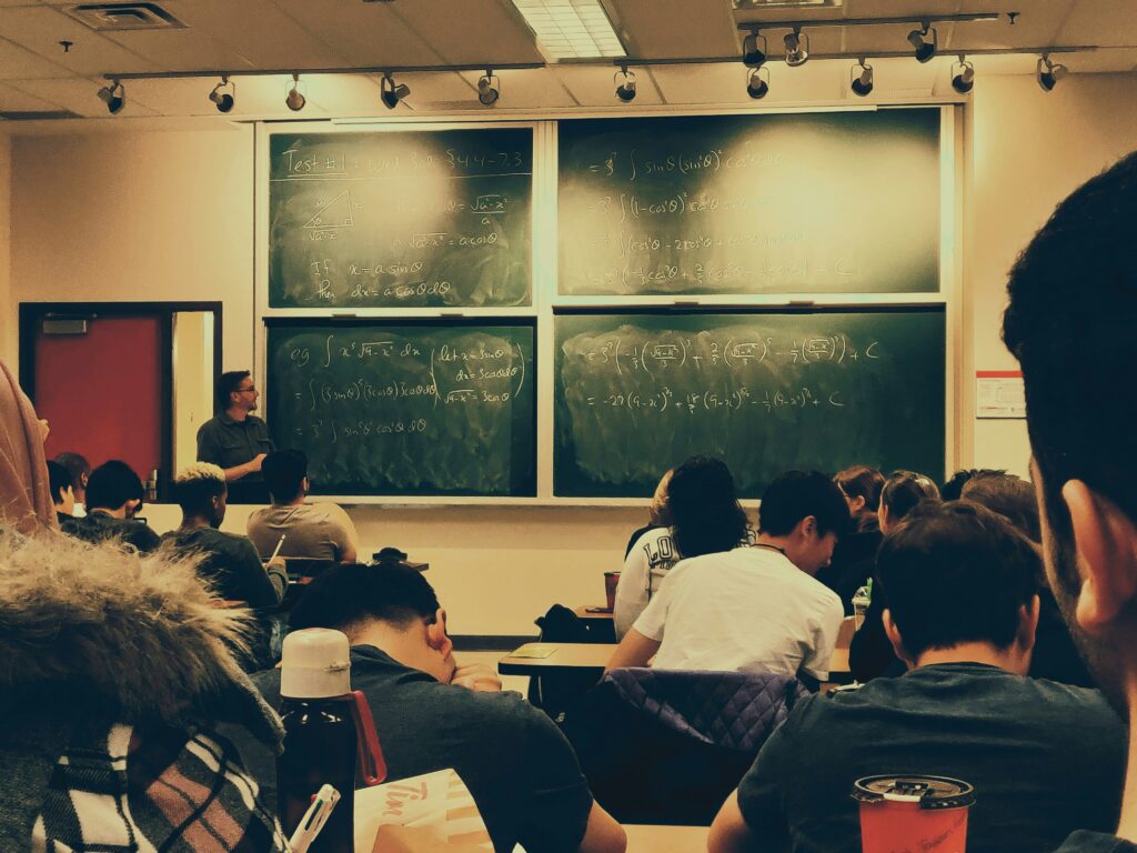 Students sitting on chairs in a classroom while the professor explains in front of a chalkboard.