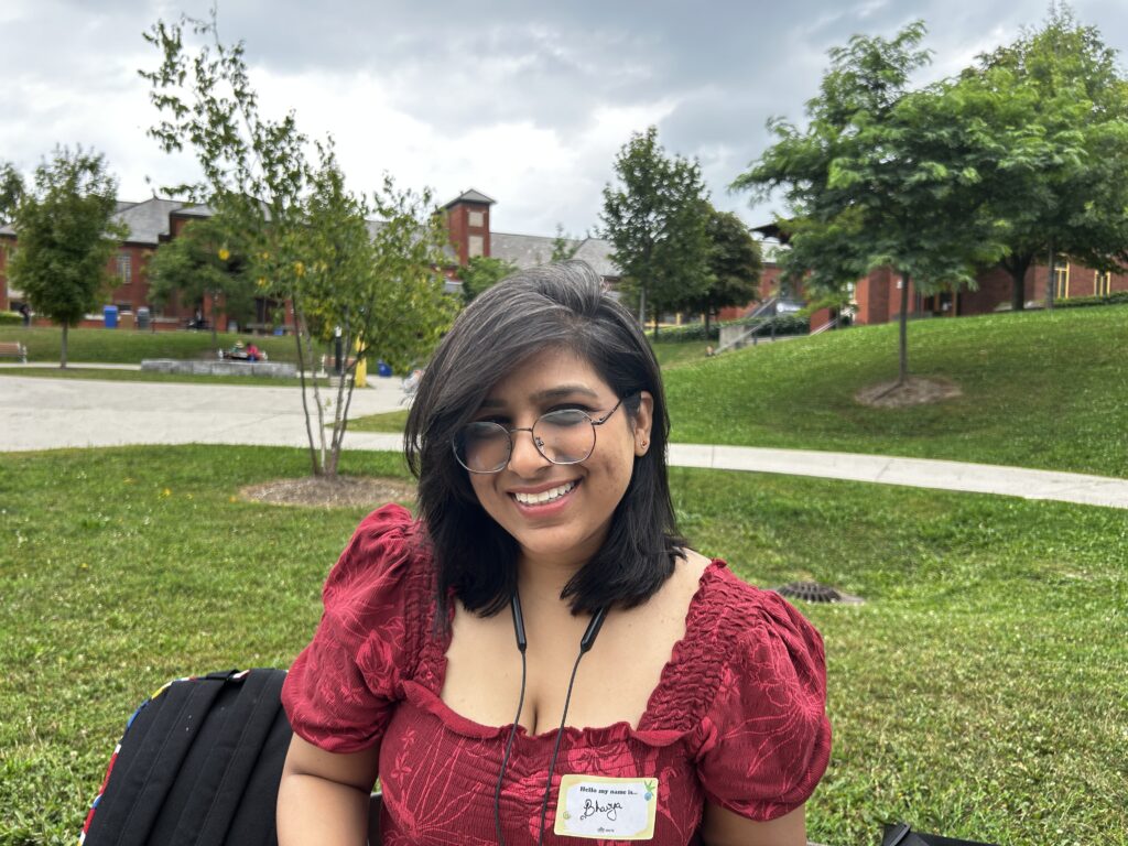 Humber student Bhavya Singh smiles as she sports a red blouse and a round pair of glasses.