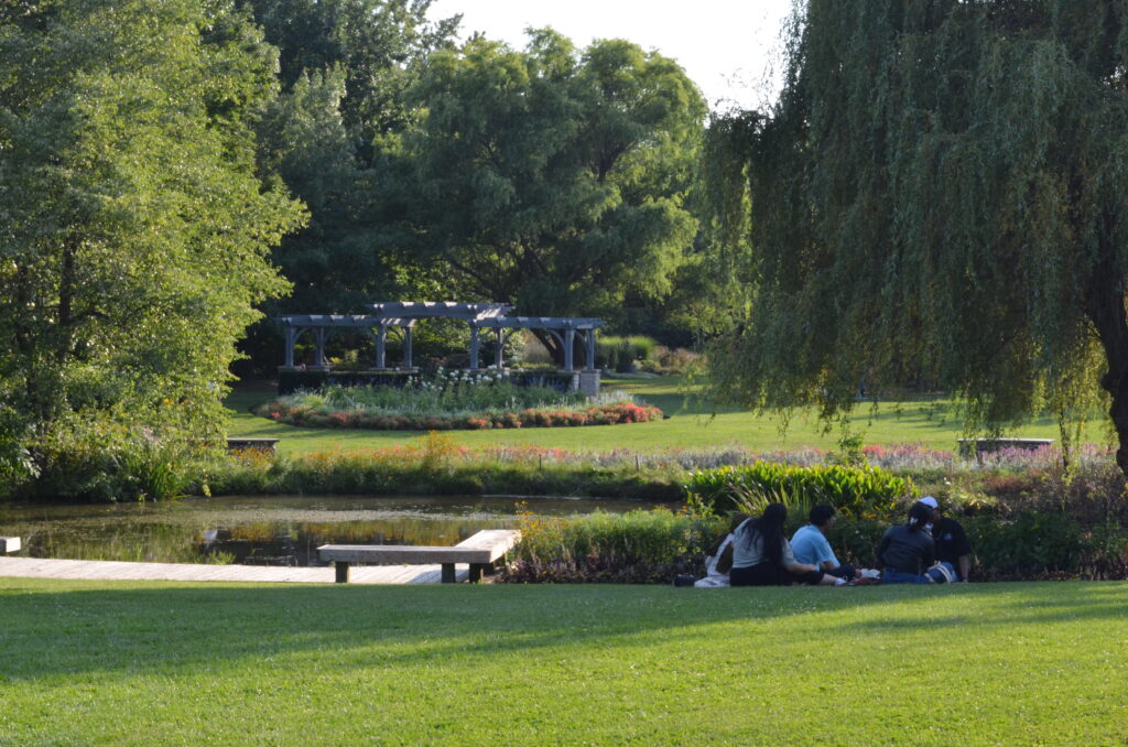 The garden is lush and green, featuring a gazebo, a pond, and a couple of friends relaxing in the grass.