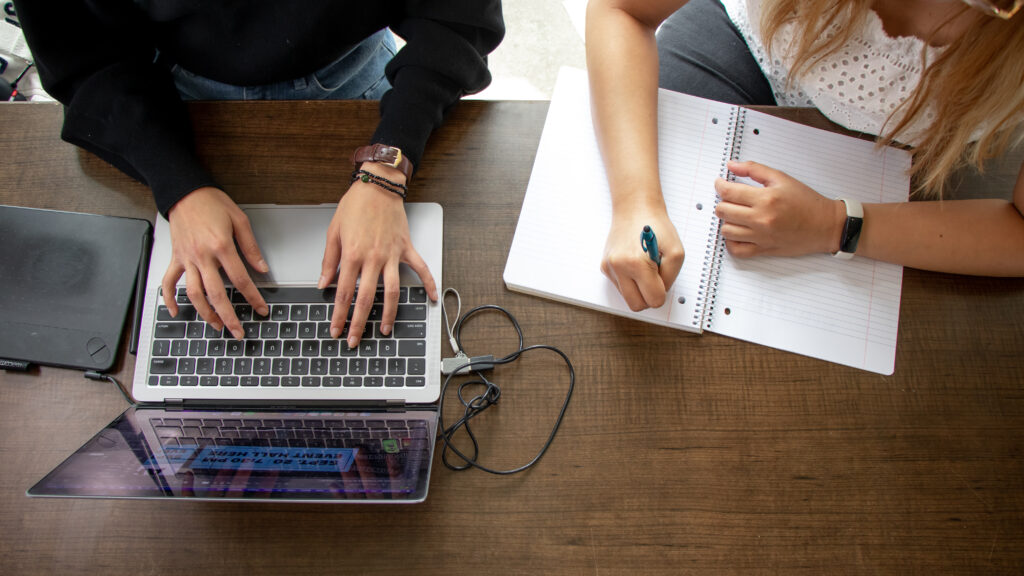 One person using a laptop and the other writing in a notebook.