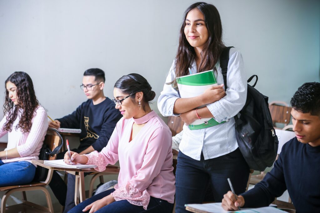 A classroom with many students doing homework and a student stood up carrying a white and green textbook and a backpack.