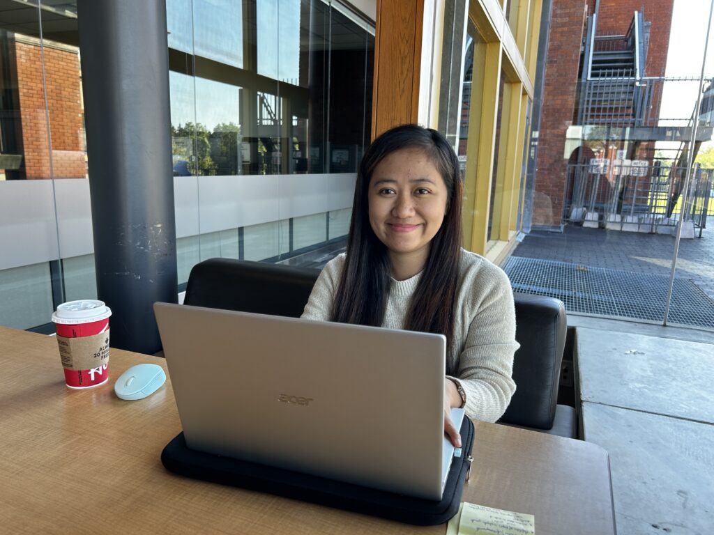 A girl smiles as she looks at the camera. She sits behind a table that contains her laptop and a cup of coffee.